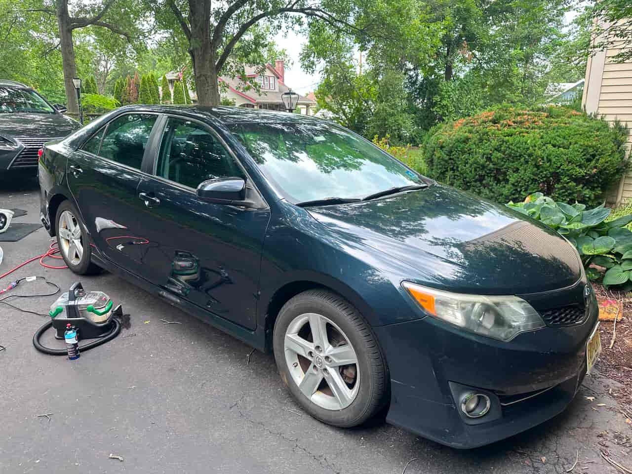 Dark blue Toyota Camry exterior BEFORE detail — dusty paint with road grime, detailing equipment set up in driveway, Flemington NJ